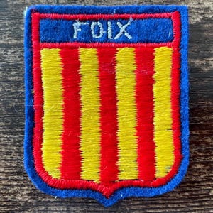 May include: A rectangular embroidered patch with a shield-shaped bottom edge. The patch features a blue border and a blue top horizontal band with the white text FOIX. The centre displays vertical red and yellow stripes throughout the design.