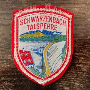 Peut inclure: Un écusson brodé en forme de bouclier avec une bordure rouge portant le texte SCHWARZENBACH TALSPERRE en rouge. Le motif représente un paysage avec un barrage, un plan d'eau, un bâtiment au toit rouge, des collines vertes et un sommet jaune.