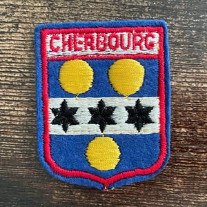 May include: Embroidered patch with a blue shield design. The patch features the word "CHERBOURG" in red letters at the top. Below are three yellow circles, three black star shapes, and a white stripe. The patch is outlined in red.