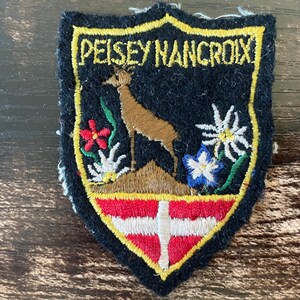 Peut inclure: Un écusson brodé en forme de blason avec un fond noir et une bordure jaune. Il présente le texte PEISEY NANCROIX en jaune, un cerf brun sur une colline, des fleurs d'edelweiss blanches, une fleur rouge et un emblème à croix rouge et blanche.