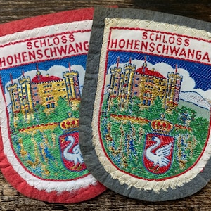 May include: Two embroidered patches featuring the Schloss Hohenschwangau castle. One patch has a red felt border, and the other has a dark gray felt border. Both display the castle, a white swan emblem on a red shield, and the text Schloss Hohenschwangau.