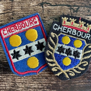 May include: Two embroidered patches featuring the coat of arms of Cherbourg. The left patch is blue with a red border, yellow circles, and black stars. The right patch is black with a gold crown, blue shield, yellow circles, and black stars.