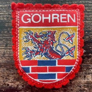 Pode incluir: Um emblema em forma de escudo com uma borda de feltro vermelho festonada. A seção superior apresenta o texto branco GOHREN em um fundo vermelho. O centro exibe um grifo vermelho em um campo amarelo e a parte inferior um padrão de tijolos.