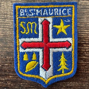 Peut inclure: Un écusson brodé bleu de forme rectangulaire portant l'inscription B&ST MAURICE en haut. Le motif comprend une croix blanche centrale avec une croix rouge superposée, une étoile jaune, les lettres SM, deux cloches jaunes, une feuille et un sapin.