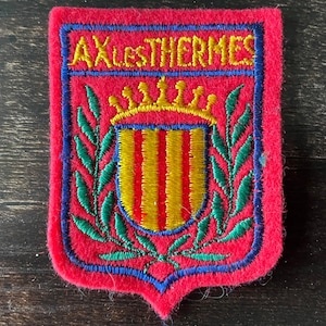 Può includere: Una toppa rettangolare in feltro rosso con uno stemma ricamato recante la scritta AX-LES-THERMES in giallo. Il disegno include uno scudo a strisce gialle e rosse sormontato da una corona gialla, fiancheggiato da rami di alloro verdi e blu.