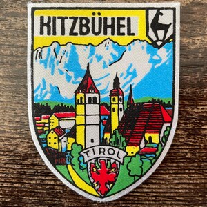 Könnte beinhalten: Ein schildförmiger Aufnäher mit dem Text KITZBÜHEL in Schwarz auf gelbem Grund. Das Motiv zeigt eine Stadt mit Kirche, rot gedeckten Häusern und schneebedeckten Bergen. Unten befindet sich ein kleines rotes Adler-Emblem.