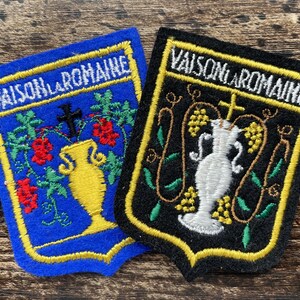 May include: Two shield-shaped embroidered patches featuring the text VAISON-LA-ROMAINE. One patch is blue with a yellow vase, red flowers, and a black cross. The other patch is black with a white vase, yellow grapes, and a yellow cross.