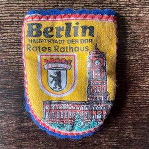 May include: A yellow, blue, and red Berlin souvenir patch. The patch features the word "Berlin" in black, the text "HAUPTSTADT DER DDR Rotes Rathaus", a bear emblem, and a depiction of a building with a clock tower.