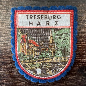 May include: A vintage patch with a blue scalloped border and a rectangular top section with the text "TRESEBURG HARZ". The patch features a colorful illustration of a town with buildings and a church.