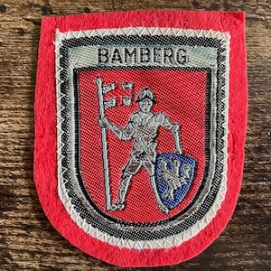 May include: A red felt patch featuring an embroidered shield design with the text BAMBERG at the top. The centre depicts a figure in armour holding a flag and a blue shield with a white lion emblem, bordered by black, white, and silver stitching.