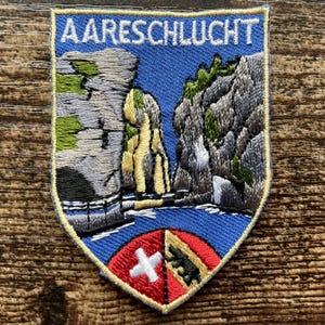 Könnte beinhalten: Ein schildförmiger Aufnäher mit der Aufschrift AARESCHLUCHT in weißen Buchstaben. Das Motiv zeigt einen blauen Fluss zwischen grauen Felsklippen mit grünen Akzenten. Am unteren Rand befindet sich ein kleines Wappen mit einem weißen Kreuz.