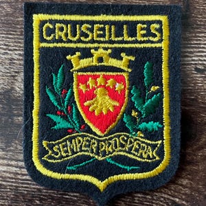 May include: A black embroidered patch featuring the text CRUSEILLES at the top and SEMPER PROSPERA on a banner at the bottom. The centre displays a red shield with a yellow emblem and crown, flanked by green leafy branches with yellow accents.