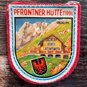 Könnte beinhalten: Ein schildförmiger Aufnäher mit rotem Rand und weißer Stickerei. Er zeigt eine Berglandschaft mit einem Steinhaus, den Text PFRONTNER HÜTTE TIROL, die Höhenangabe 1800 m und ein kleines schwarzes Wappen mit einem roten Adler-Emblem.