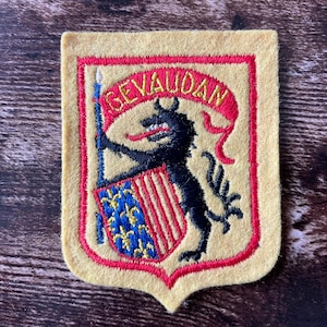 Può includere: Toppa ricamata con un disegno a scudo. La toppa presenta un lupo nero che tiene una lancia, uno stendardo rosso con la scritta "GEVAUDAN" e uno scudo con gigli blu e oro. La toppa ha uno sfondo giallo con bordi rossi.