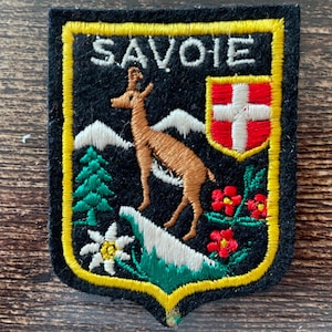 May include: A black embroidered patch with a yellow border featuring the text SAVOIE in white. The design includes a brown chamois, white mountain peaks, a green pine tree, a white edelweiss flower, red flowers, and a red shield with a white cross.