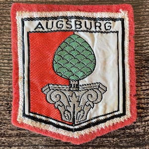 Könnte beinhalten: Ein rechteckiger gestickter Aufnäher mit rotem Rand und dem Wort AUGSBURG in schwarzer Schrift oben. In der Mitte befindet sich ein Wappenschild in Rot und Weiß mit einem grünen Pinienzapfen auf einem grauen steinernen Kapitell.
