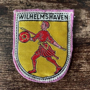 Op de afbeelding: Geborduurde patch met de tekst "WILHELMSHAVEN" boven een rode figuur met een schild en speer op een gele achtergrond. De patch heeft een roze rand en witte stiksels.