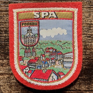 Op de afbeelding: Een schildvormige geborduurde patch met een rode vilten rand. Het midden toont een landschap van de stad Spa, inclusief het Pouhon-gebouw, met de tekst SPA bovenaan en POUHON op een kleiner wapenschild. Het ontwerp gebruikt blauw, groen en rood.