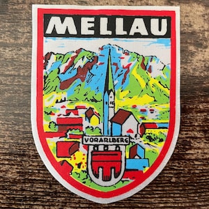 Op de afbeelding: Een schildvormige stoffen patch met een kleurrijke illustratie van het stadje Mellau in Vorarlberg, Oostenrijk. Het ontwerp bevat een centrale kerktoren, bergtoppen en gebouwen, met de tekst MELLAU en VORARLBERG erop gedrukt.