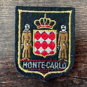 Puede incluir: Parche bordado negro con borde dorado. El parche presenta un escudo con una corona, dos figuras y un patrón rojo y blanco. El texto "MONTE-CARLO" está bordado en rojo.