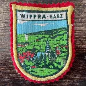 Könnte beinhalten: Ein schildförmiger, bestickter Aufnäher mit einer Landschaft von Wippra Harz, einer Kirche, grünen Hügeln und blauem Himmel. Der Aufnäher hat ein weißes oberes Banner mit dem schwarzen Text WIPPRA HARZ, einen gelben Rand und einen roten Wellenrand.
