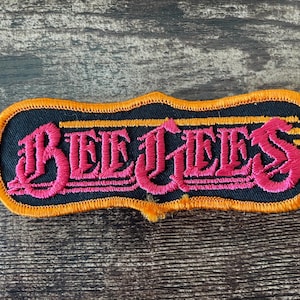 Può includere: Toppa ricamata con la scritta "BEE JEEZ" in rosa, con lettere stilizzate. La toppa ha uno sfondo nero con un bordo arancione. La toppa è rettangolare con la parte superiore e inferiore ondulata.