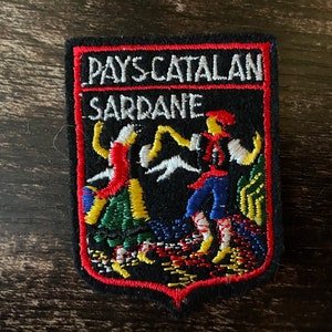 May include: Embroidered patch with the text "PAYS CATALAN SARDANE" in white on a black background. The patch features two figures dancing in colorful attire, with red, yellow, green, and blue details. The patch has a red border.
