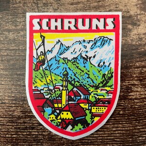 Op de afbeelding: Een kleurrijke patch met het woord "SCHRUNS" bovenaan. De patch toont een berglandschap met een skilift, gebouwen en bergen op de achtergrond. De patch heeft een rode rand.