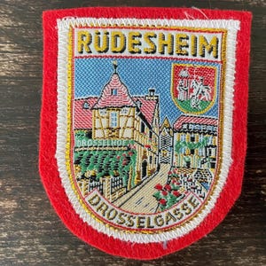 May include: A rectangular embroidered patch with a red felt border featuring a detailed scene of Rudesheim and Drosselgasse. The design includes a half-timbered building, a small coat of arms, and the text Rudesheim and Drosselgasse in gold thread.
