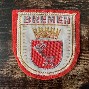 May include: A shield-shaped embroidered patch featuring a red border and a white center. The text BREMEN is stitched in silver at the top. Below, a gold crown sits above a red shield containing a silver key, all outlined with gold thread.