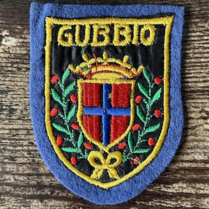May include: A blue felt patch featuring an embroidered crest with the text GUBBIO in yellow. The design includes a central red shield with a blue cross, topped by a yellow crown, surrounded by green laurel branches with red berries and a yellow bow.