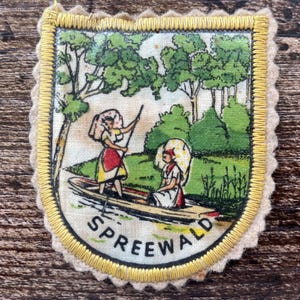 Könnte beinhalten: Ein rechteckiger Stoffaufnäher mit gewelltem Rand und gelber Stickkante. In der Mitte befindet sich eine gedruckte Illustration von zwei Personen in einem kleinen Boot auf einem Fluss, umgeben von grünen Bäumen. Das Wort SPREEWALD ist schwarz aufgedruckt.