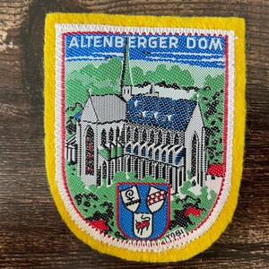 May include: A rectangular embroidered patch with a yellow border featuring a detailed depiction of the Altenberger Dom cathedral. The design includes a blue sky, green trees, and a shield with three smaller crests at the bottom, all in vibrant thread.