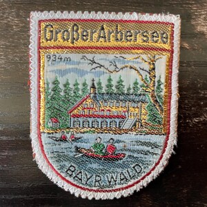 May include: A rectangular embroidered patch featuring a landscape scene of the Großer Arbersee lake with a building, trees, and people in a red boat. The text Großer Arbersee, 934m, and BAYR WALD is stitched in black thread on a gold and white background.