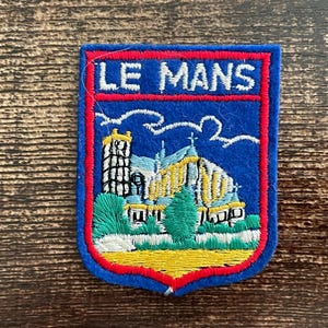 Op de afbeelding: Een schildvormige geborduurde patch met een blauwe achtergrond en een rode rand. De tekst LE MANS is bovenaan in het wit geborduurd. Daaronder staat een wit en geel gebouw met een hoge toren afgebeeld boven groen gebladerte en een gele basis.