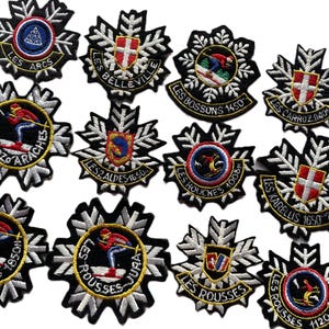 May include: A collection of embroidered patches with various designs and text. The patches feature ski resort names like "Les Arcs" and "Les Menuires", along with skier illustrations and shield emblems. The patches are black, white, red, and yellow.