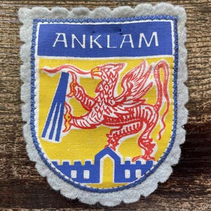 May include: A shield-shaped fabric patch with a scalloped light grey felt border. The centre features a yellow field with a red griffin holding a blue object, above a blue castle wall. The top blue banner displays the white text ANKLAM.
