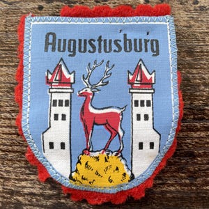 Puede incluir: Un parche de tela en forma de escudo con fondo azul claro y el texto Augustusburg en negro. El diseño incluye dos torres blancas con techos rojos flanqueando un ciervo rojo sobre un montículo amarillo, bordeado por fieltro rojo.