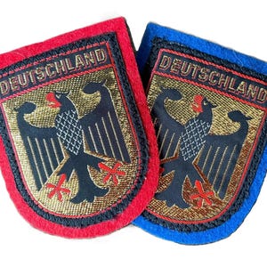 Può includere: Due toppe ricamate a forma di scudo con la parola DEUTSCHLAND in lettere maiuscole nere nella parte superiore. Ogni toppa mostra un'aquila nera con accenti rossi su sfondo dorato, bordata da una spessa cornice nera e feltro rosso o blu.
