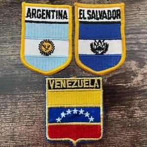 Può includere: Tre toppe ricamate a forma di scudo con le bandiere di Argentina, El Salvador e Venezuela. Ogni toppa ha un bordo giallo e riporta il nome del paese in nero nella parte superiore, insieme al rispettivo emblema nazionale o alle stelle.