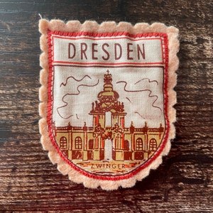 May include: A fabric patch with a scalloped edge. The patch features the word "DRESDEN" at the top, with an illustration of a building below. The word "ZWINGER" is at the bottom of the patch, which is in a shield shape.