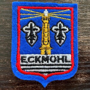 Könnte beinhalten: Ein schildförmiger, bestickter Aufnäher mit blauem Filzhintergrund und rotem Rand. Er zeigt einen gelben Leuchtturm mit strahlenden Lichtkegeln, zwei schwarz-weiße Lilien-Symbole und den Text ECKMOHL in Schwarz auf einem weißen Balken.