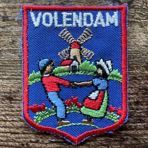Könnte beinhalten: Ein rechteckiger gestickter Aufnäher mit blauem Hintergrund und rotem Rand. Der Text VOLENDAM ist oben in Weiß gestickt. Das Design zeigt zwei Figuren, die sich vor einer Windmühle, grünem Gras und roten Blumen an den Händen halten.