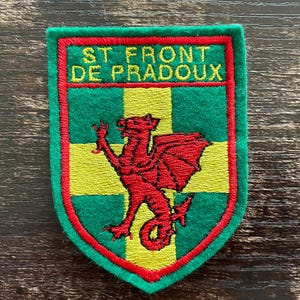Könnte beinhalten: Ein schildförmiger, bestickter Aufnäher mit grünem Hintergrund, einem gelben Kreuz und einem roten Drachen in der Mitte. Der obere Bereich enthält den gelb gestickten Text ST FRONT DE PRADOUX, umrandet von einem dicken roten Rand.