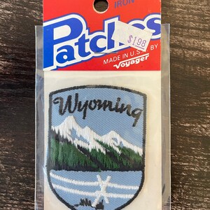 Può includere: Una toppa ricamata termoadesiva a forma di scudo con cielo azzurro, montagne bianche innevate, pini verdi e una staccionata bianca. La parola Wyoming è ricamata in corsivo nero. La confezione riporta le scritte Patches, Iron-on e Voyager.
