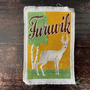 Puede incluir: Parche bordado con la palabra "Tururik" en escritura burdeos sobre un ciervo blanco con cuernos. El fondo presenta un árbol verde y un campo amarillo, con árboles blancos y verdes a la izquierda. El parche tiene un borde deshilachado blanco.
