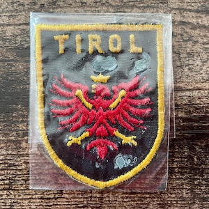 Pode incluir: Um patch em forma de escudo com a palavra "TIROL" em letras amarelas no topo. O patch apresenta um emblema de águia vermelha com detalhes amarelos sobre um fundo preto, emoldurado por uma borda amarela. O patch está em uma manga de plástico transparente.