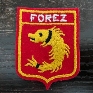 Peut inclure: Un écusson brodé rouge de forme rectangulaire avec une base en pointe. Il présente une créature jaune brodée ressemblant à une crevette avec des détails noirs et le texte blanc brodé FOREZ en haut, le tout bordé d'un fil jaune.