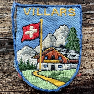 May include: A rectangular embroidered patch with a light blue background featuring the word VILLARS in yellow thread. The design depicts a red Swiss flag, white snow-capped mountains, a green pine tree, and a chalet with a yellow path.