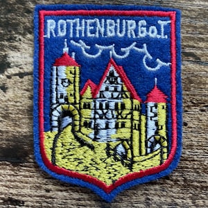 May include: A shield-shaped embroidered patch featuring a blue background with a red border. The design depicts a yellow and white building with red roofs and the text ROTHENBURG o.T. in white thread at the top, with a white line graphic above.